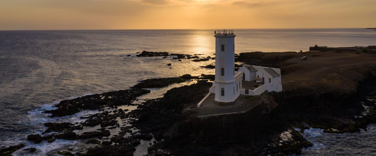 Um farol numa costa rochosa com o sol se pondo ao fundo.