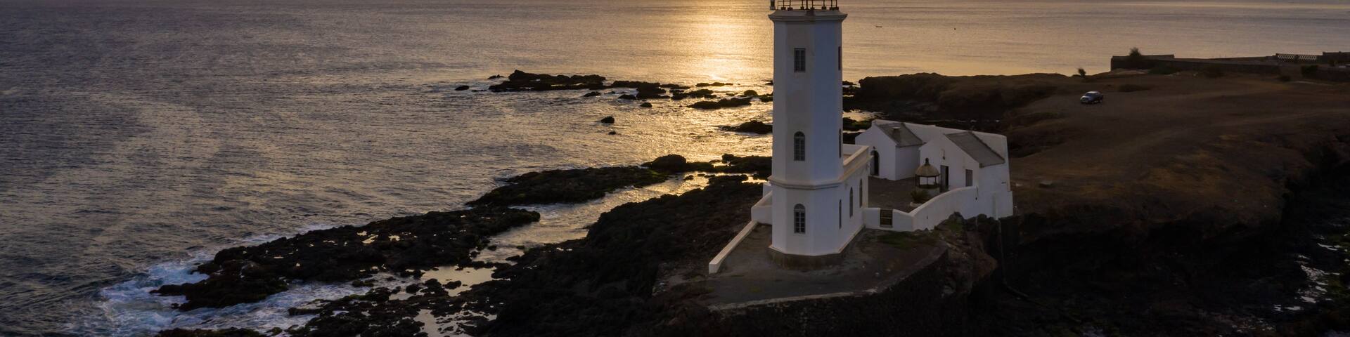 A lighthouse on a rocky coastline with the sun setting in the background.