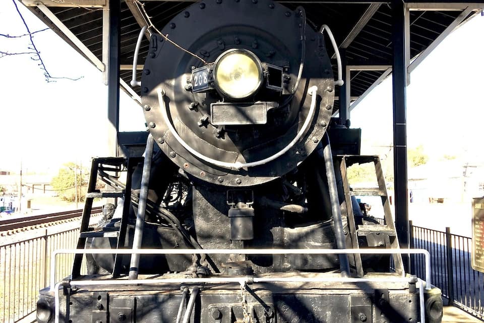 Una locomotora de vapor estacionada, con un farol delantero destacado y el número 208, ubicada bajo una plataforma cubierta con techo metálico.