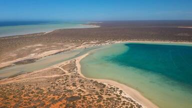 Vista aérea de uma área costeira com uma grande baía, praias de areia e vegetação rala.