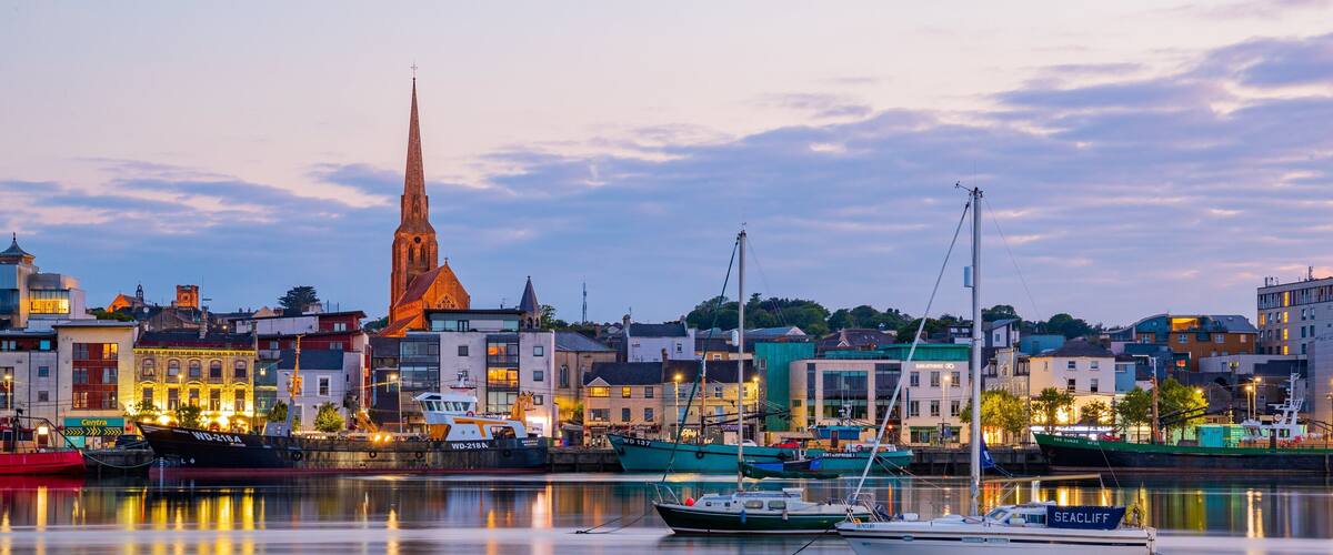 A waterfront cityscape with a prominent church spire, several boats docked along the quay, and a mix of historic and modern buildings.