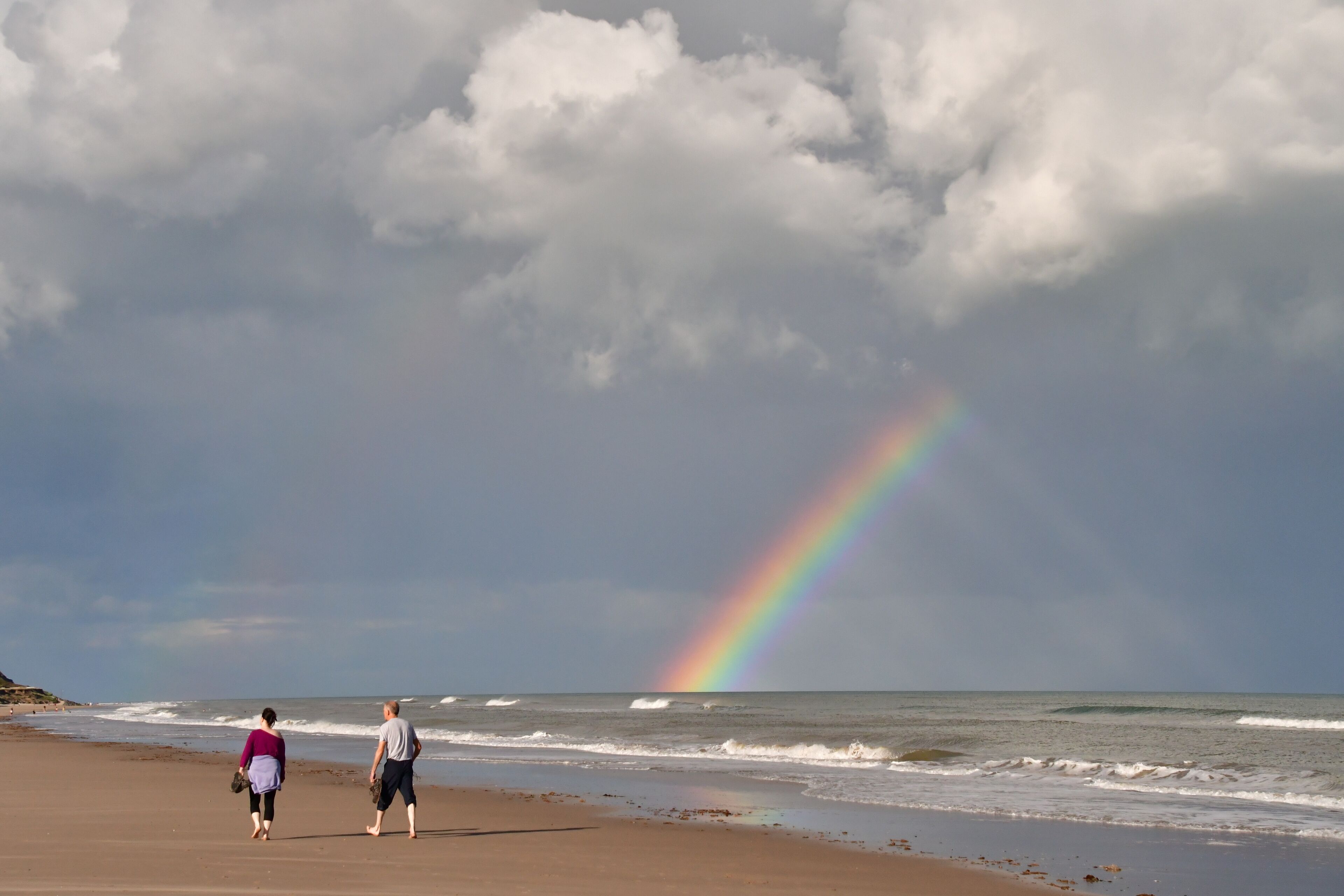 Una playa con un arcoíris, dos personas caminando y olas.