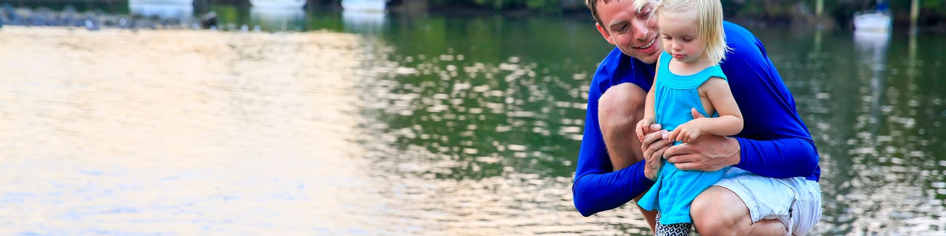 Un hombre y un niño sobre una roca junto a un lago tranquilo con barcos al fondo.