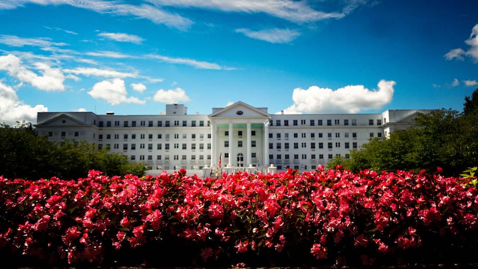 A large white building with a central entrance, surrounded by lush green trees and a vibrant red flower bed in the foreground.