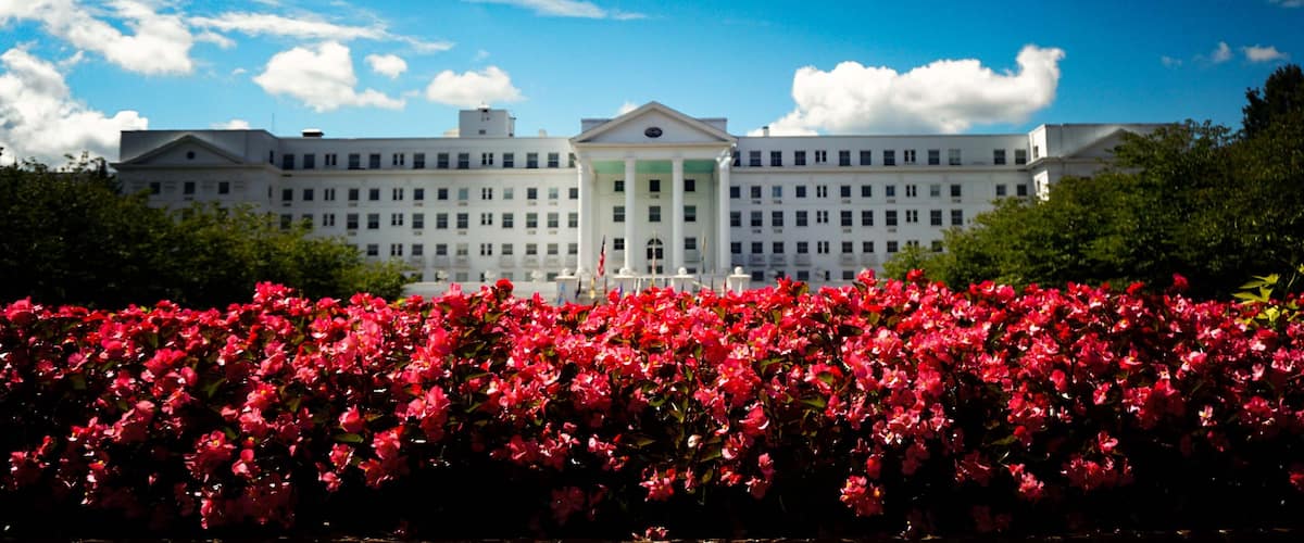 A large white building with a central entrance, surrounded by lush green trees and a vibrant red flower bed in the foreground.