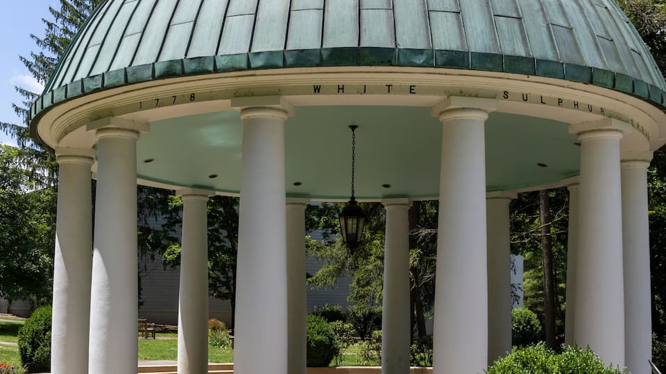 A gazebo with a green dome and white columns, surrounded by red flowers and green shrubs.