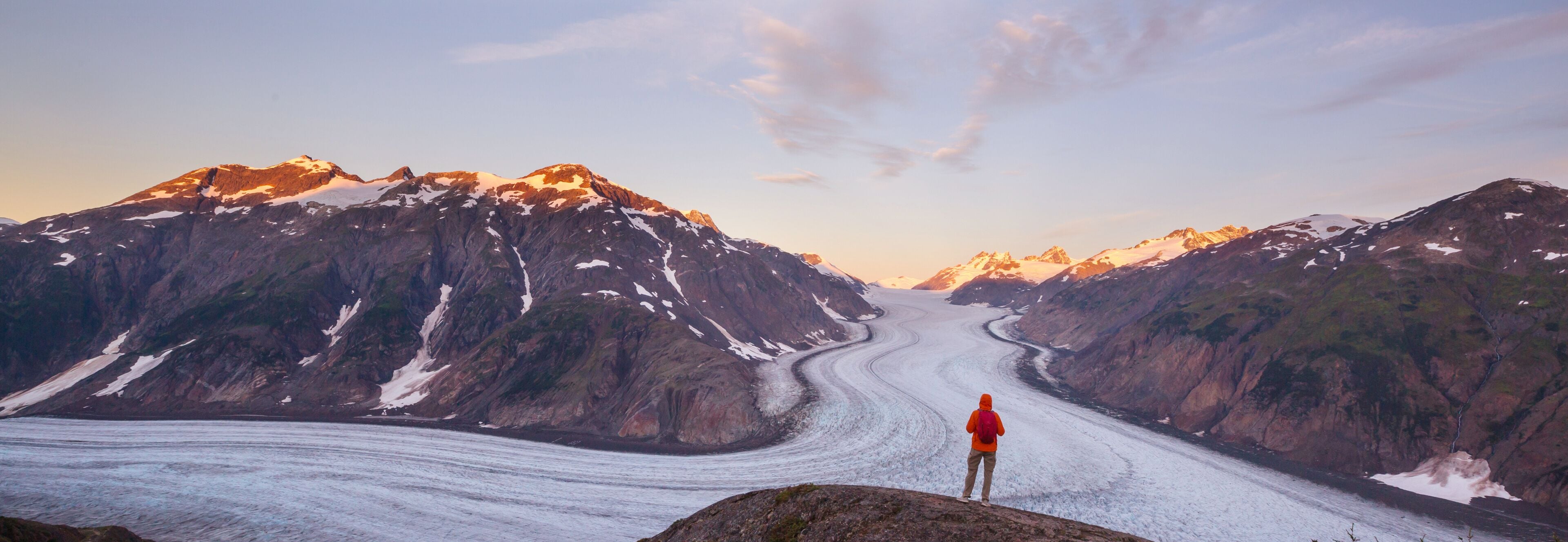 Um glaciar serpenteando por uma paisagem montanhosa, com uma pessoa em uma rocha.
