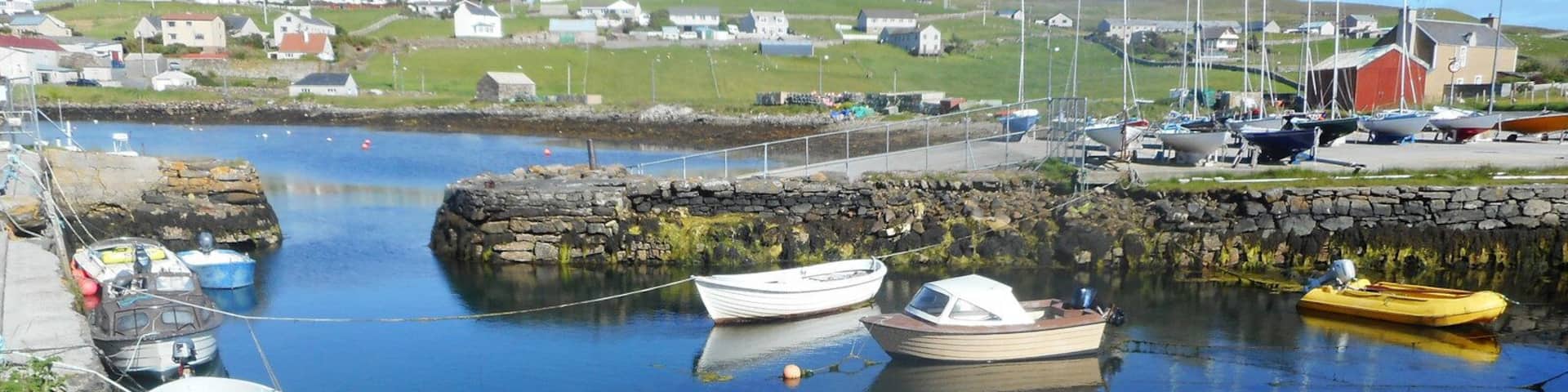 A harbor with several boats docked, a stone breakwater, and houses in the background.