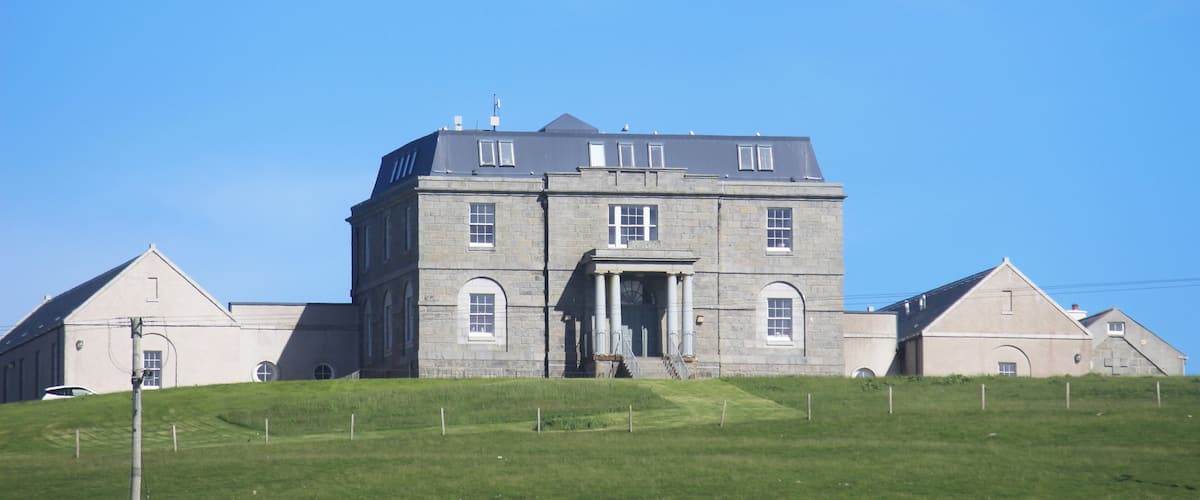 A large stone building with a central entrance and two wings, surrounded by a grassy hill.