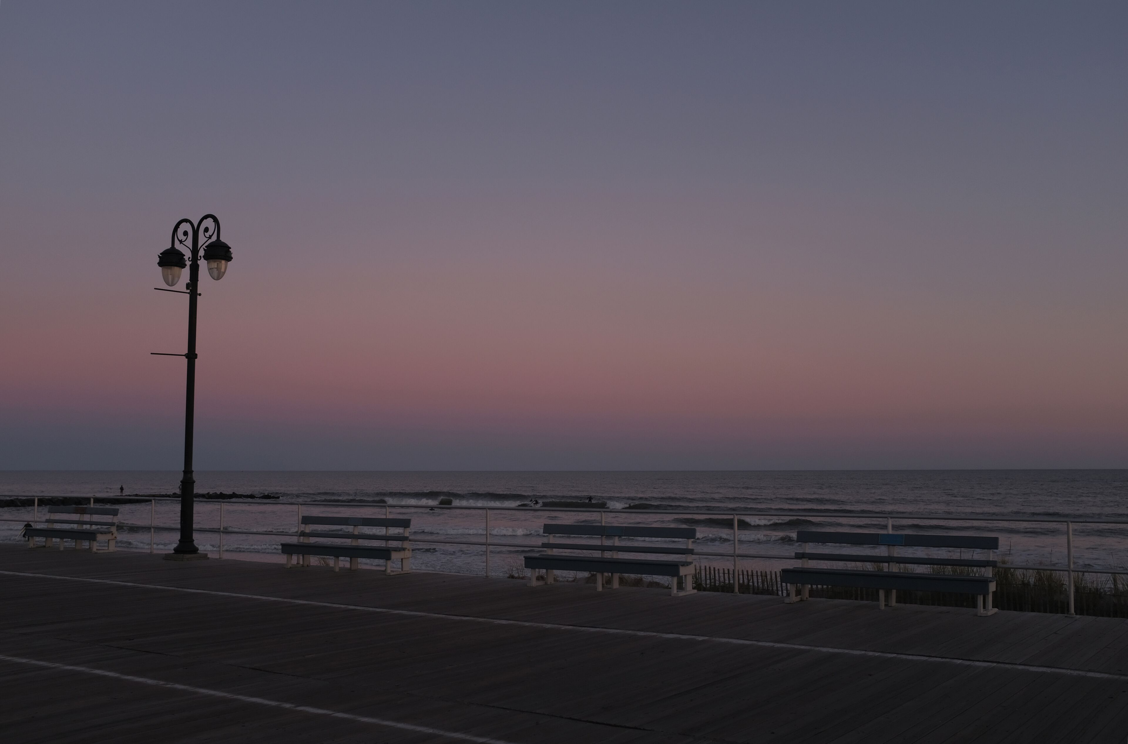 A sunset over the ocean with a boardwalk, benches, and a street lamp.