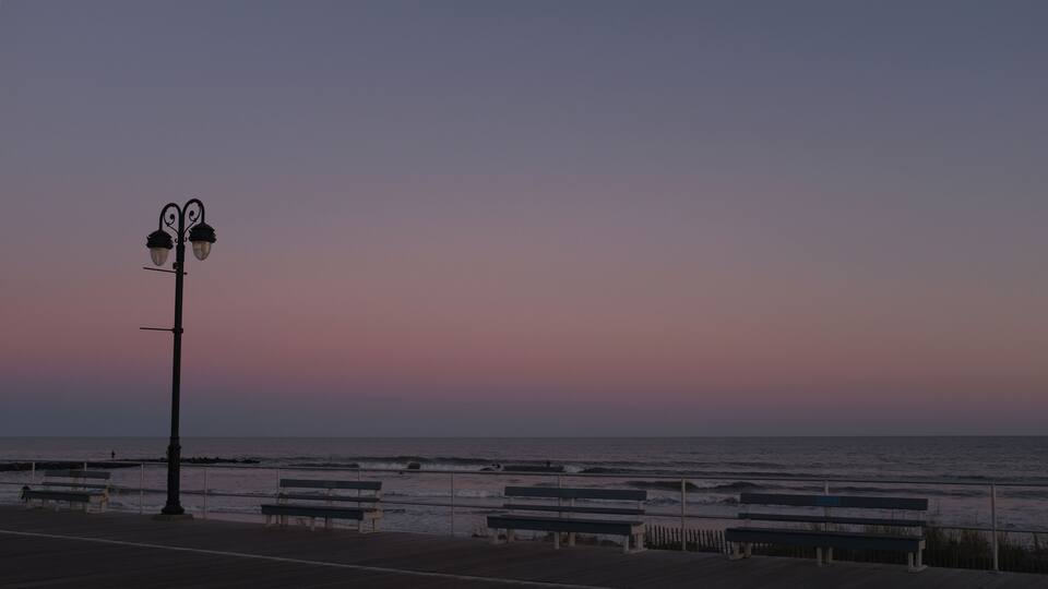 A sunset over the ocean with a boardwalk, benches, and a street lamp.