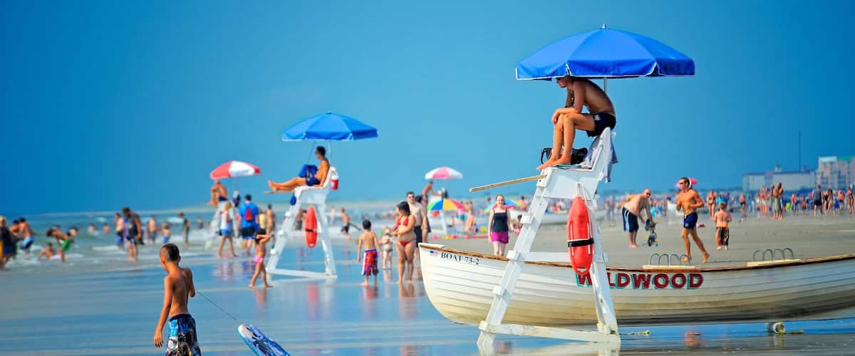 A lifeguard tower with a lifebuoy, a beach with people, and a boat named 'BOAT 73-2' with 'WILDWOOD' written on it.