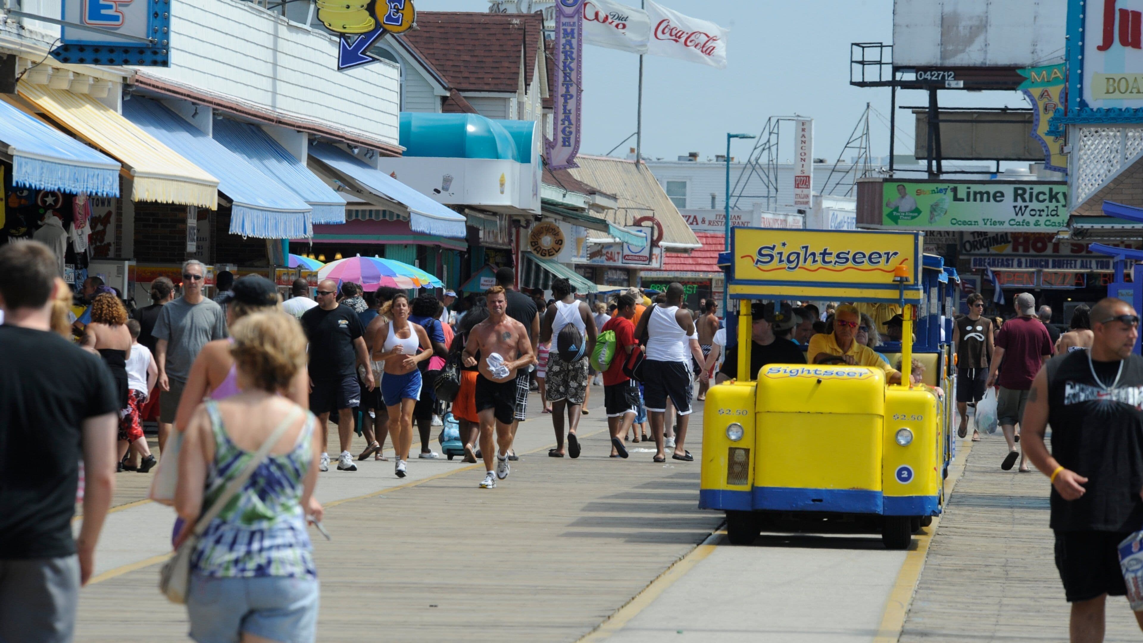 A crowded boardwalk with a yellow sightseeing trolley, shops, and a Coca-Cola sign.