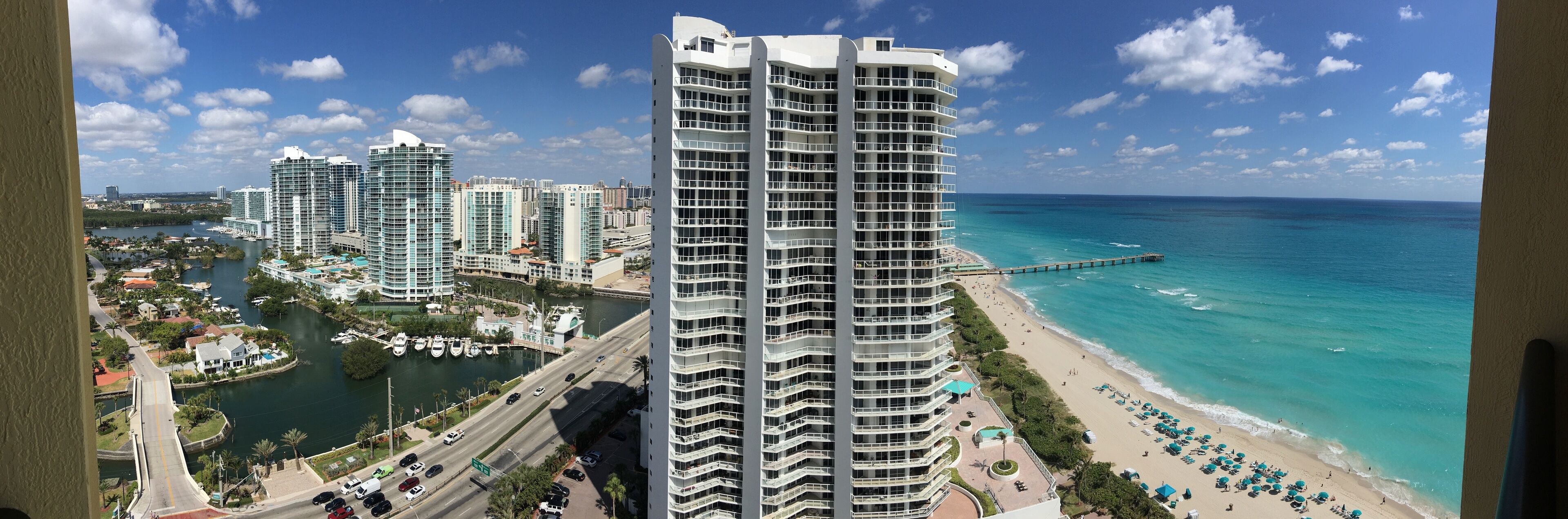 A coastal cityscape with high-rise buildings, a marina, and a beach with umbrellas.