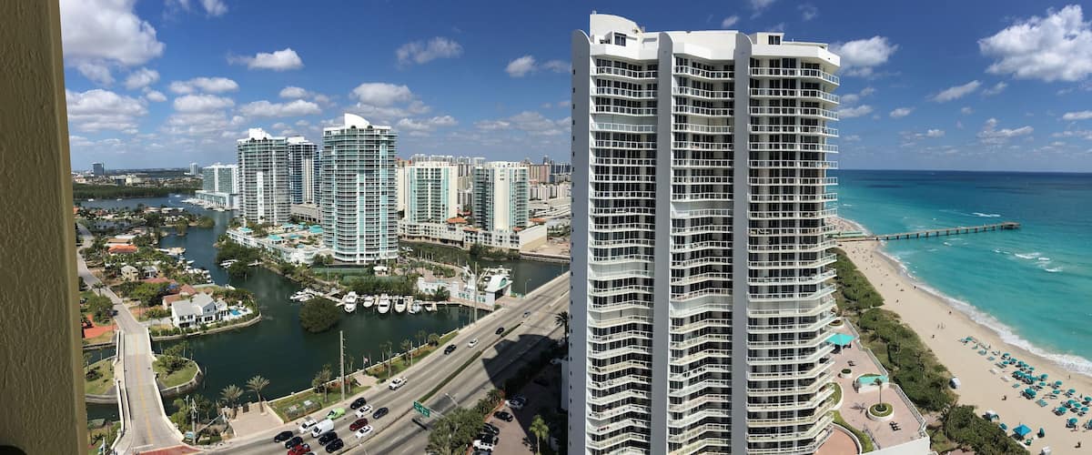 A coastal cityscape with high-rise buildings, a marina, and a beach with umbrellas.