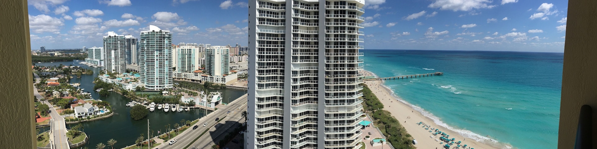 A coastal cityscape with high-rise buildings, a marina, and a beach with umbrellas.