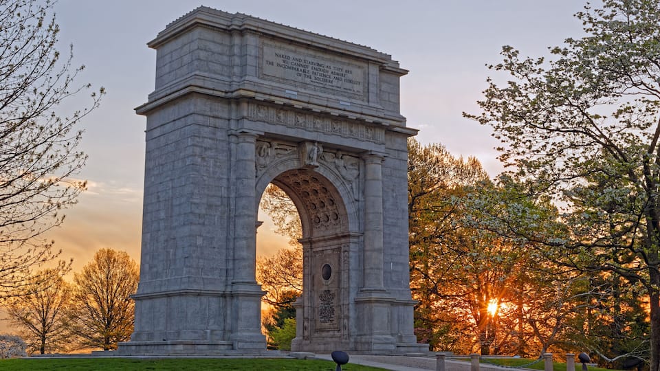 A large stone arch monument with inscriptions, surrounded by trees and a sunset in the background.