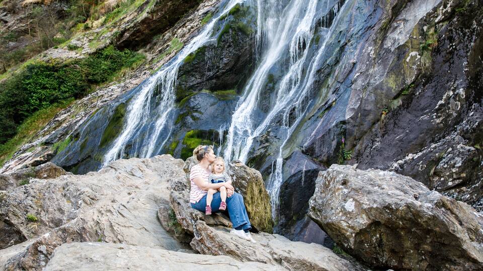 A waterfall cascading over rocks with a person and child sitting on a rock in front.
