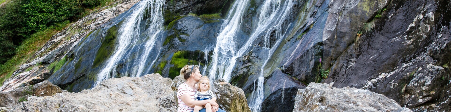 Una cascada fluye sobre rocas, con una persona y un niño sentados sobre una roca al frente.