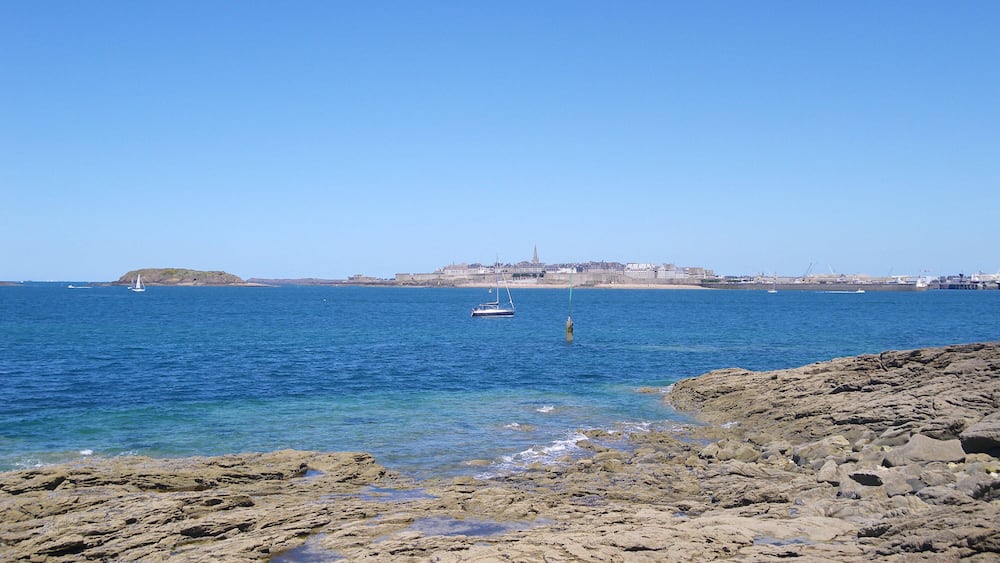 Eine felsige Küstenlinie mit klarem, blauem Wasser, einer Segelyacht und einer fernen Stadtlandschaft mit Gebäuden und einem Turm.