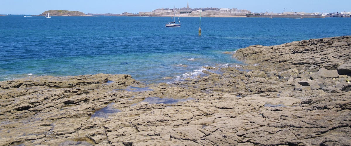A rocky shoreline with clear blue water, a sailboat, and a distant cityscape with buildings and a tower.