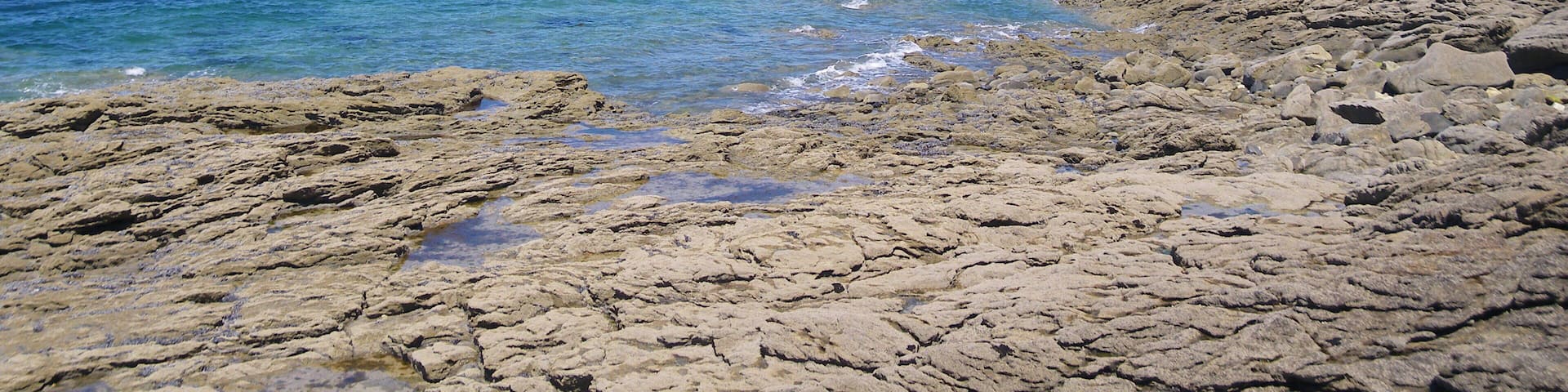 A rocky shoreline with clear blue water, a sailboat, and a distant cityscape with buildings and a tower.