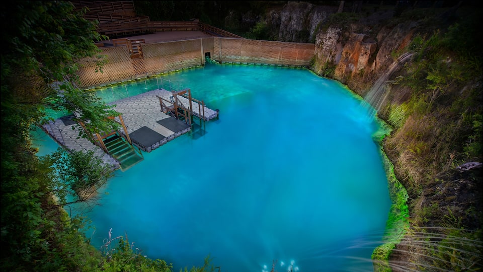 A turquoise pool with a wooden deck and steps, surrounded by greenery and a rocky cliff.