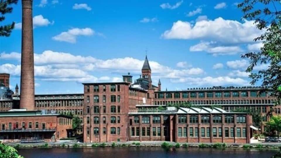 A large industrial building with a tall chimney, a domed structure, and a river in the foreground.