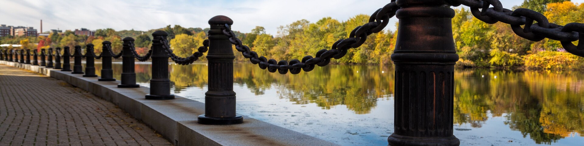 Un paseo marítimo con una fila de boletas negras unidas por cadenas, un camino pavimentado y una masa de agua tranquila que refleja los árboles y el cielo circundantes.