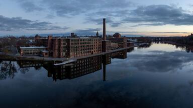 Un río refleja un edificio industrial de ladrillo con una chimenea alta, rodeado de árboles y un cielo nublado.