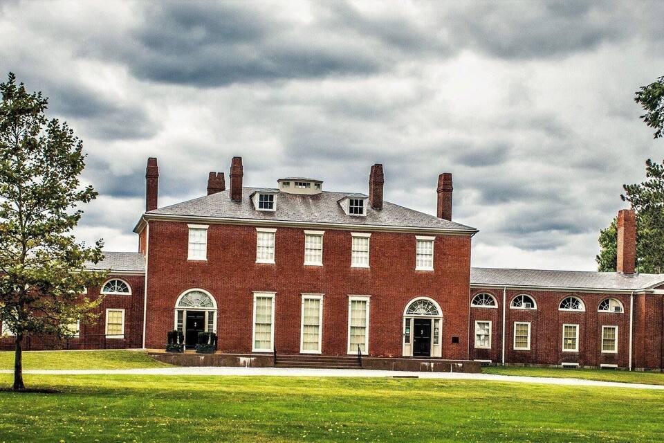 A large brick building with arched windows and a central entrance, surrounded by a well-maintained lawn and trees.