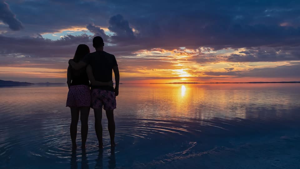 A couple standing on a reflective water surface at sunset.