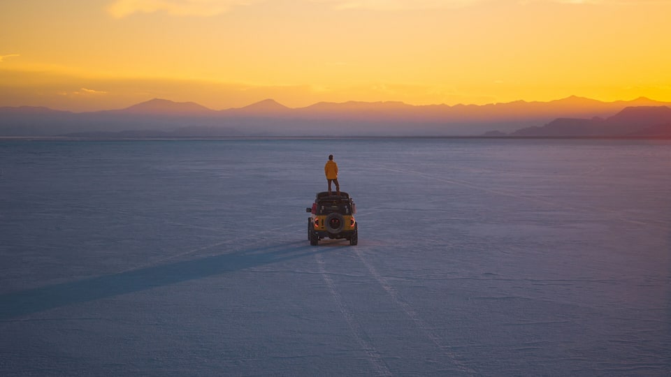 A person standing on a vehicle in a vast salt flat with mountains in the background during sunset.