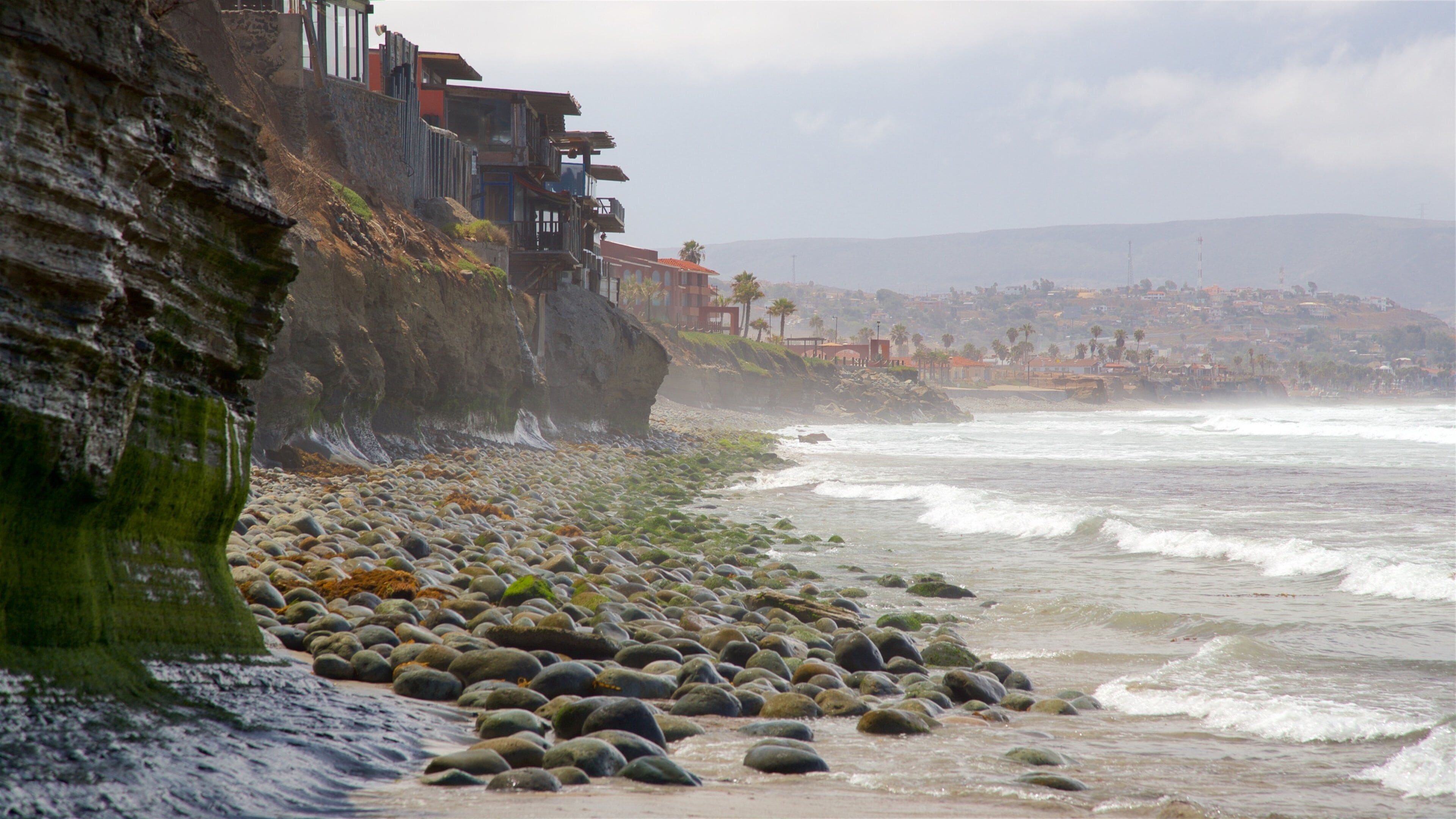 Una playa rocosa con olas rompiendo en la orilla, un acantilado con edificios y un pueblo a lo lejos.