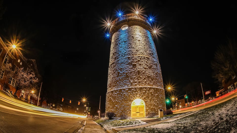 A tall, illuminated tower with a circular window and a door, surrounded by snow and streetlights.