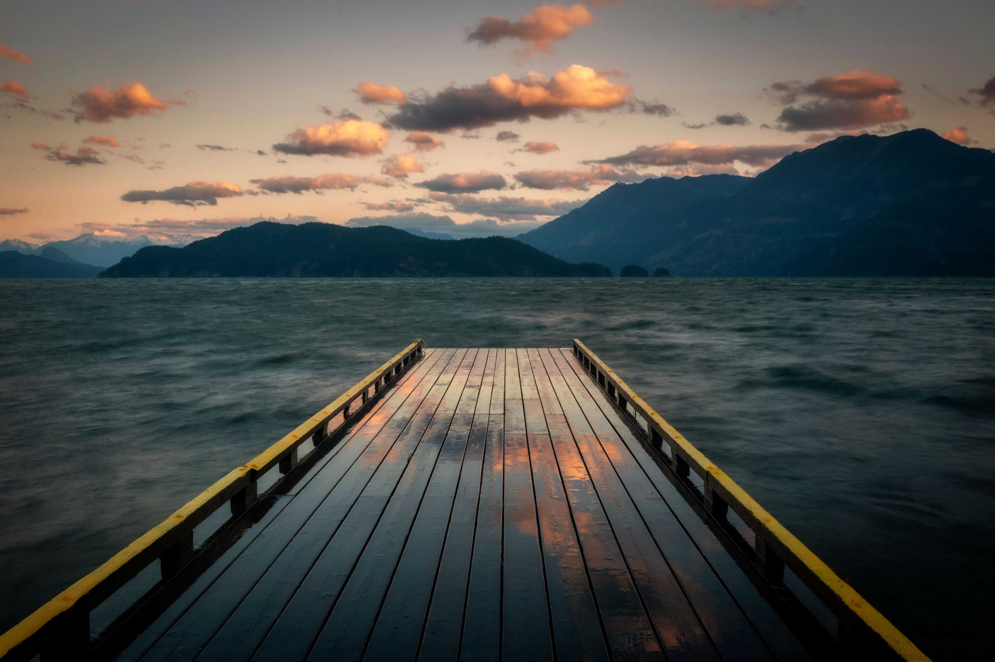 A wooden pier extending into calm water with mountains in the background.