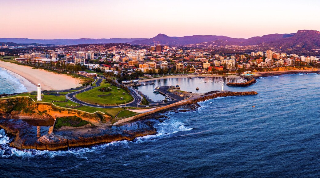 An aerial view of a coastal city with a prominent lighthouse, a beach, and a mountainous backdrop.