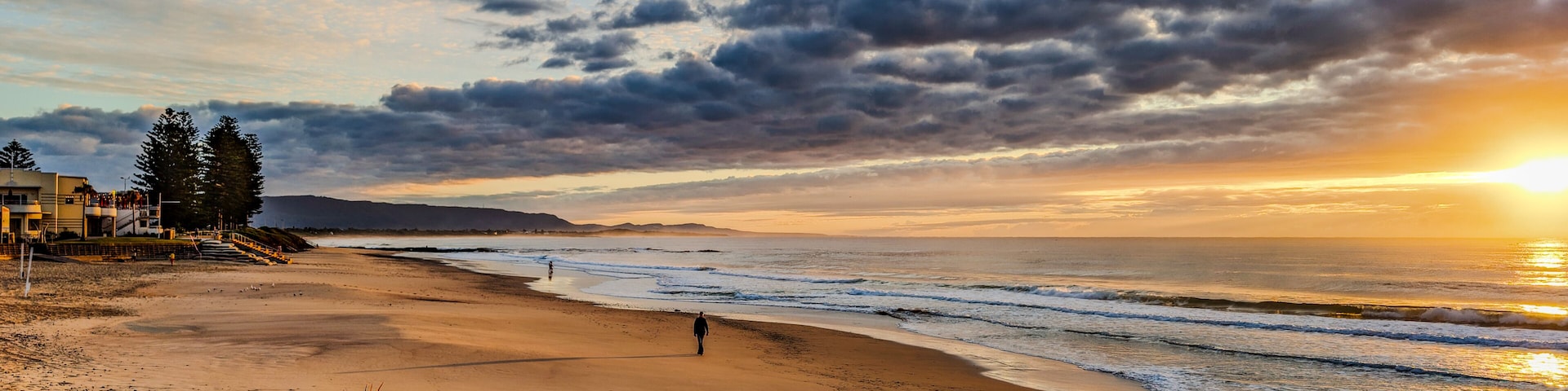 Uma praia com uma pessoa caminhando, ondas do oceano e um pôr do sol.