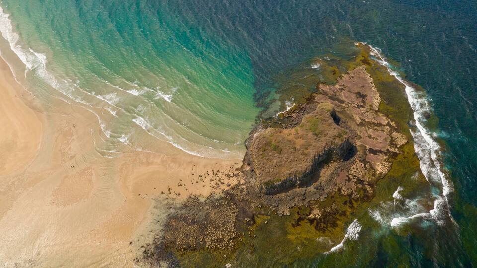A coastal landscape with a sandy beach, a rocky outcrop, and clear blue waters.