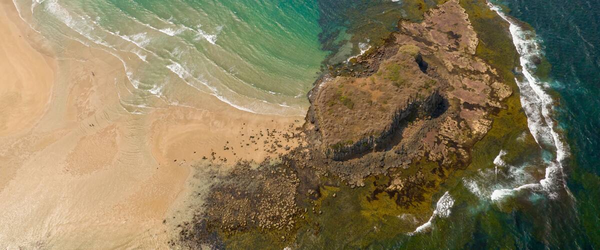 A coastal landscape with a sandy beach, a rocky outcrop, and clear blue waters.