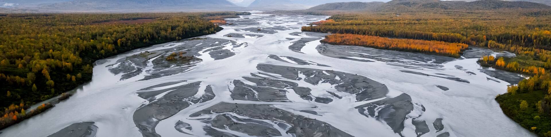 A river with ice formations surrounded by autumn-colored trees and mountains in the background.