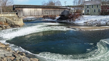 A frozen river with a wooden bridge, a stone dam, and houses in the background.
