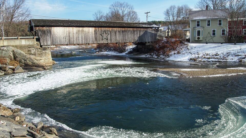 A frozen river with a wooden bridge, a stone dam, and houses in the background.