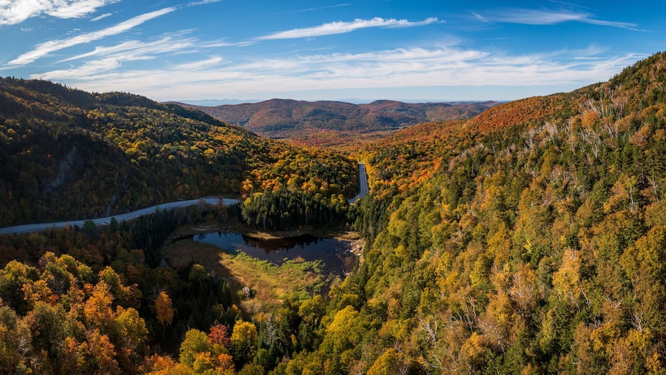 A winding road through a forested valley with a lake and distant mountains.