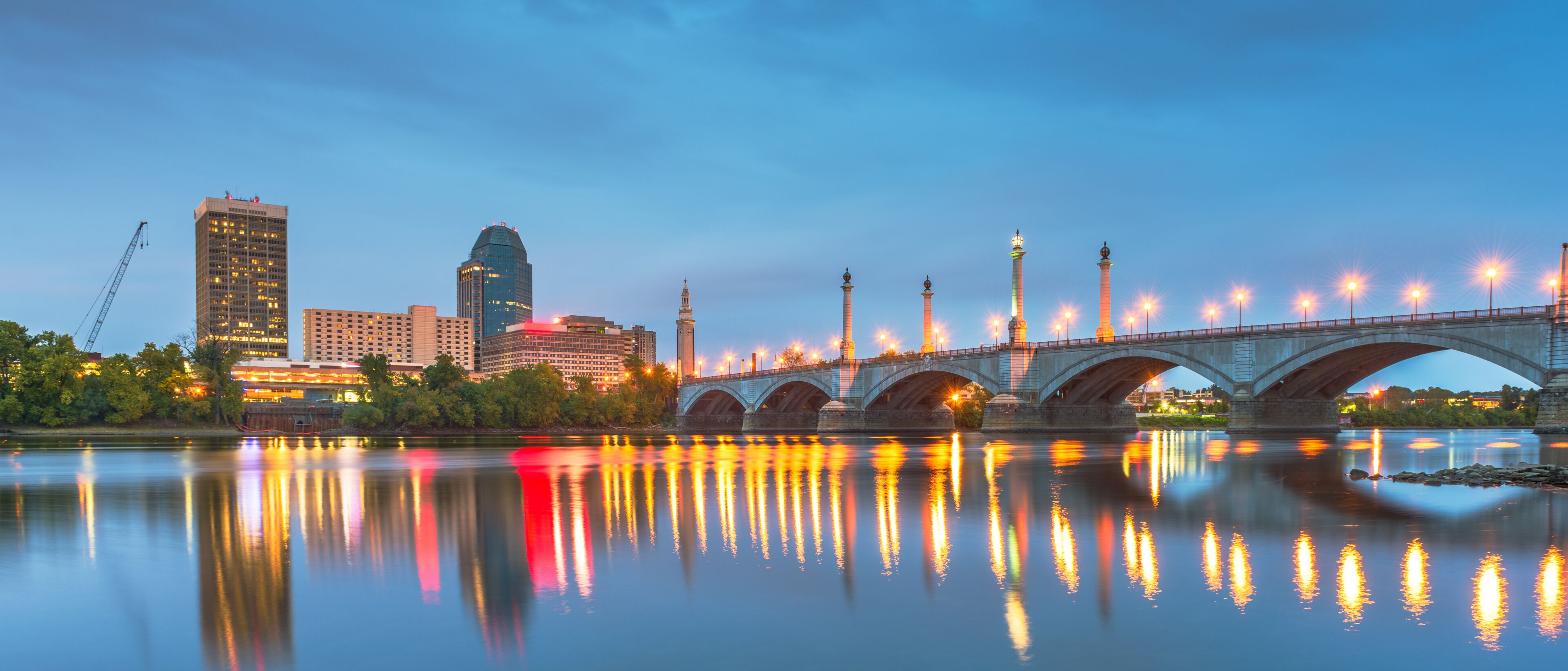 A city skyline with illuminated buildings, a bridge, and a calm river reflecting the lights.