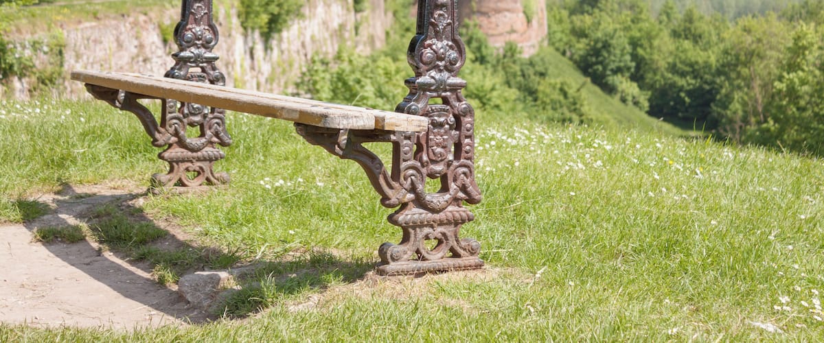 A bench with ornate metalwork in a grassy area with a view of a forested hill and a distant landscape.