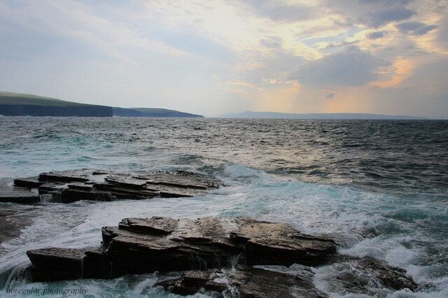 A rocky coastline with waves crashing against the rocks under a cloudy sky.