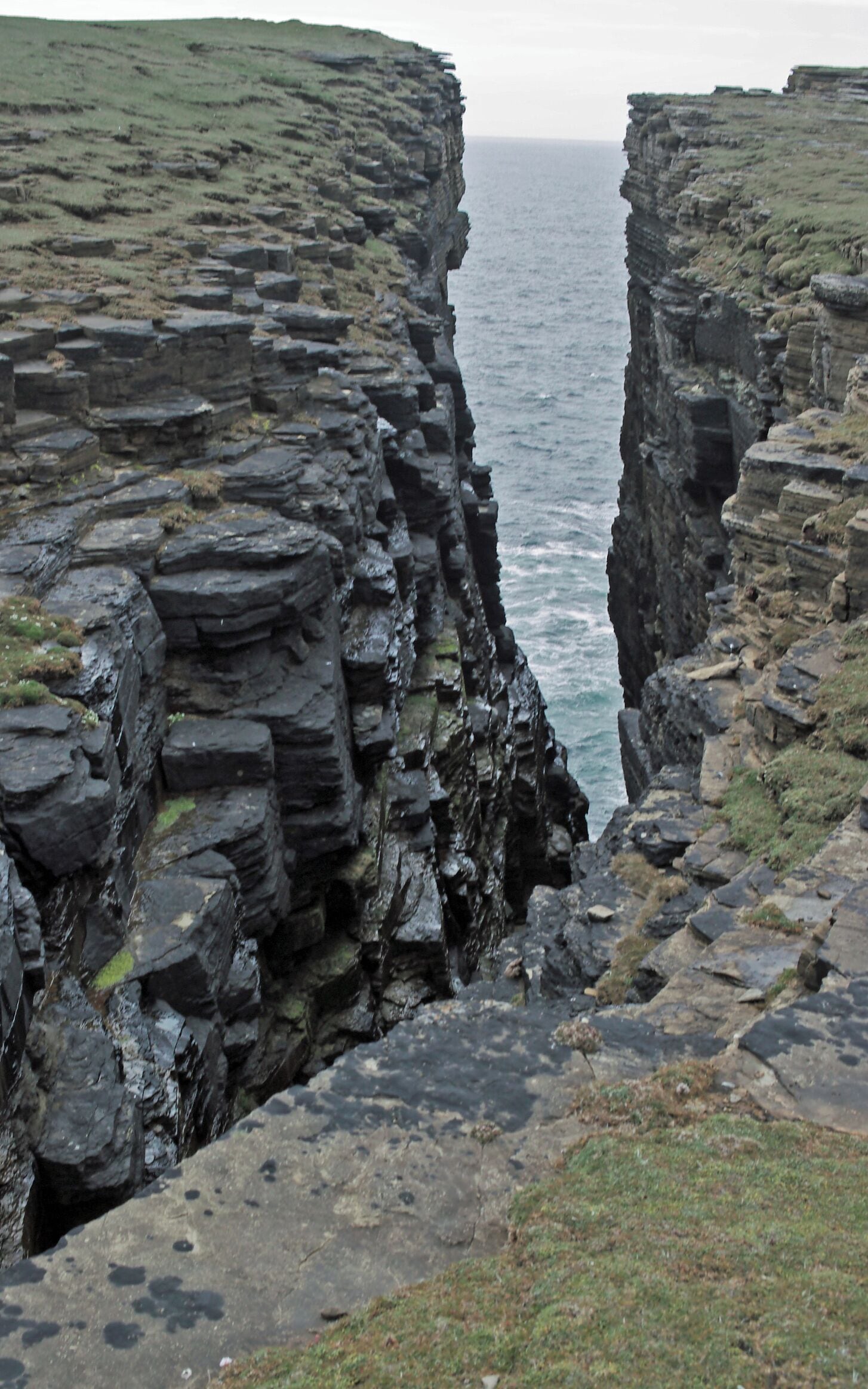 A rugged coastal cliff with layered rock formations and a narrow sea passage.