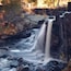 Une cascade d’eau qui dévale des rochers dans une zone boisée aux couleurs automnales.
