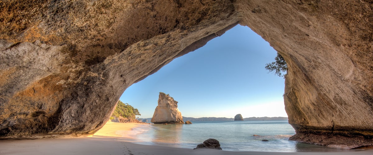 Eine natürliche Felsformation als Tor über einem Strand, mit Fußspuren, die zum Wasser führen.